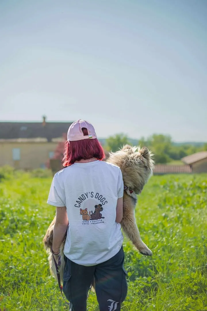 Candice, de dos, marche dans un pré ensoleillé en tenant un petit chien dans ses bras.